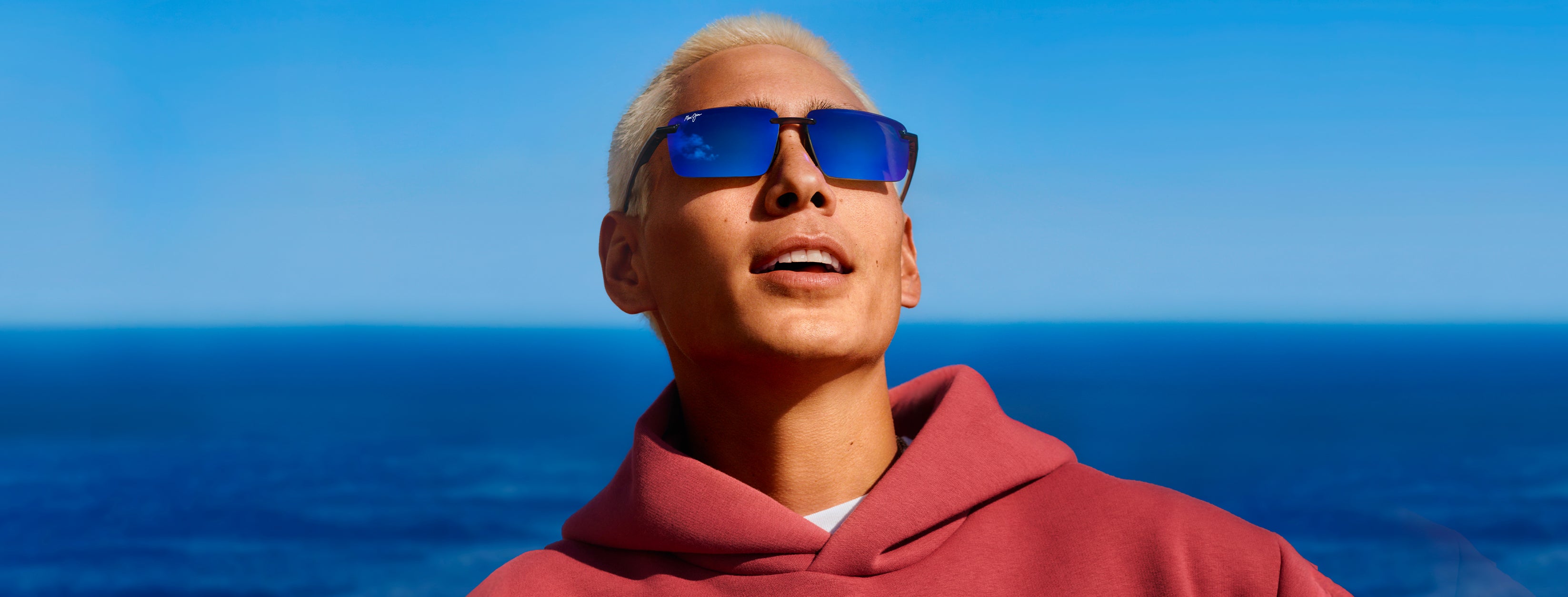Person wearing maui jim sunglasses and a red hoodie with a blue sky and ocean background