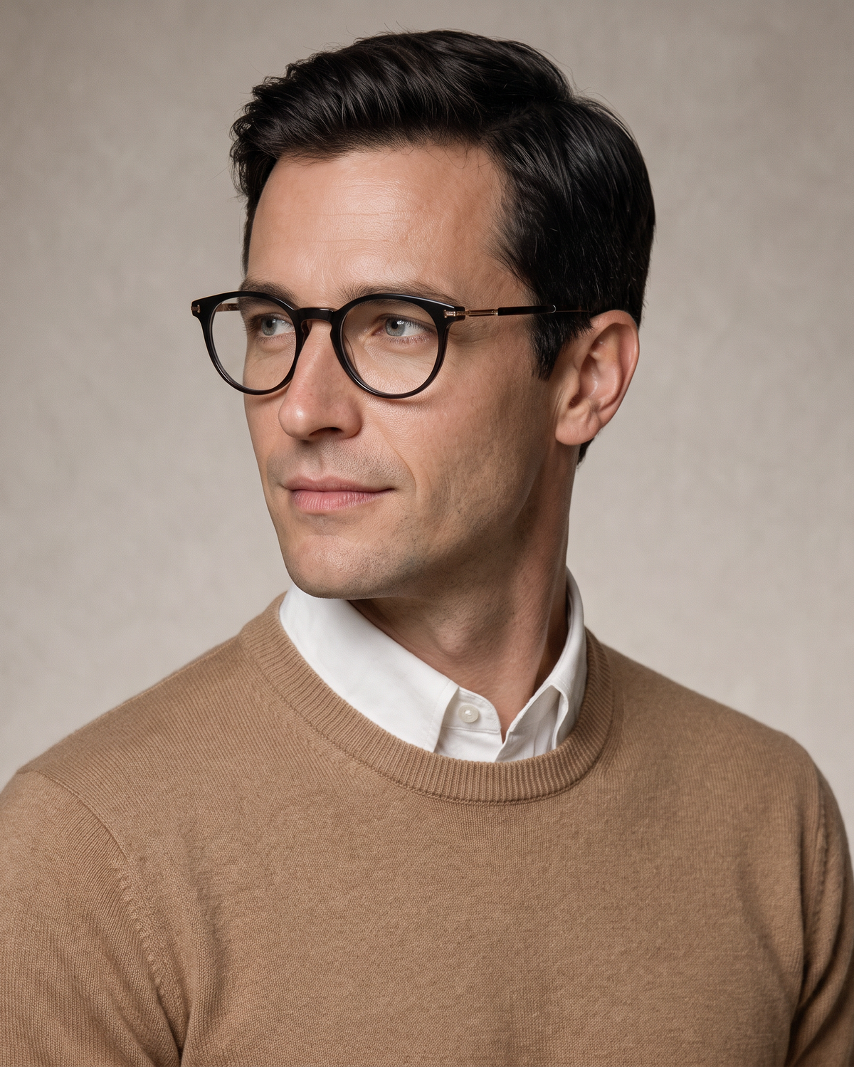 A man wearing black round-frame eyeglasses, a tan crewneck sweater, and a white collared shirt, photographed against a neutral beige background.